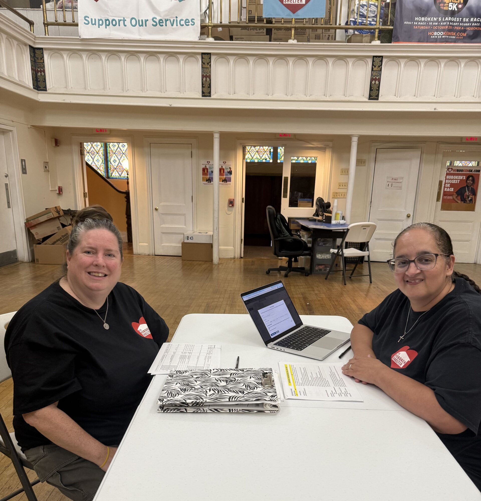 The Hoboken Shelter’s Executive Director, Jaclyn Cherubini, and Clinical Case Coordinator, Norma Rosario, smile while seated together at a table with a laptop and paperwork inside the Shelter.